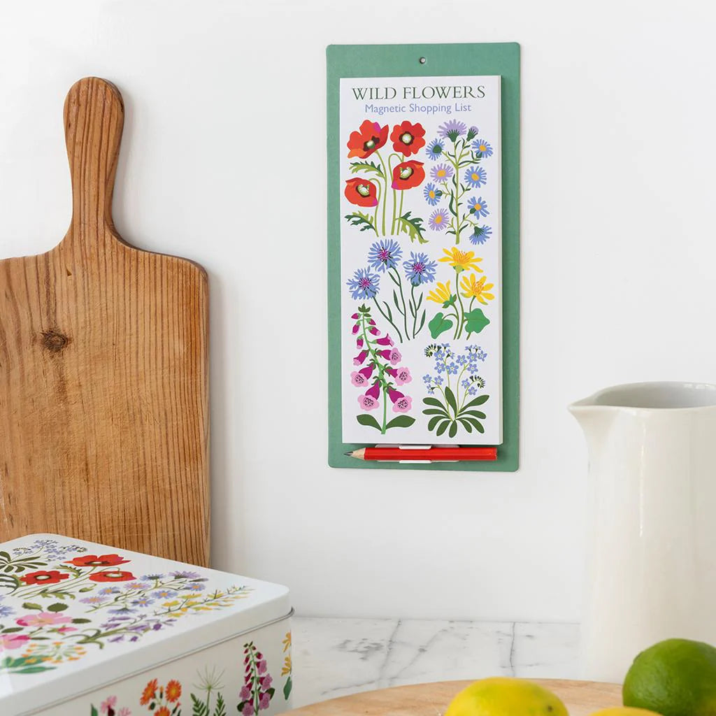 Magnetic shopping list with wildflower design on a white wall next to a wooden cutting board and floral container.
