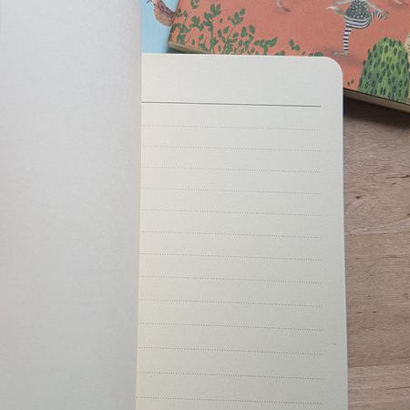 Open notebook with lined pages on a wooden surface, with two colorful notebooks in the background.
